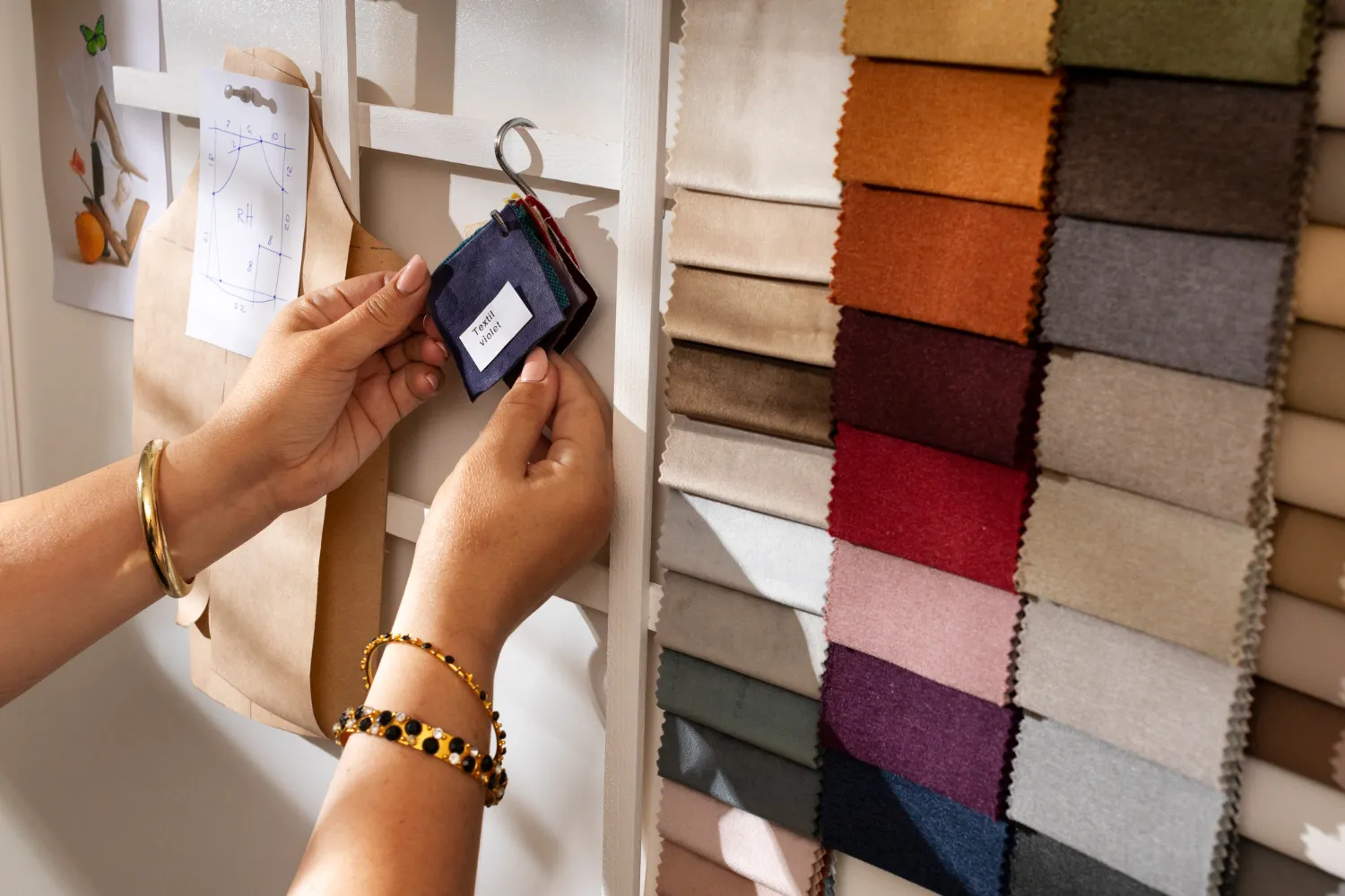 Manos sosteniendo una muestra de tela color violeta frente a una pared con una variedad de muestras de telas en diferentes colores, junto a un boceto de diseño de moda.